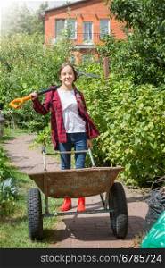 Portrait of happy young girl posing with wheelbarrow and shovel in garden at sunny day