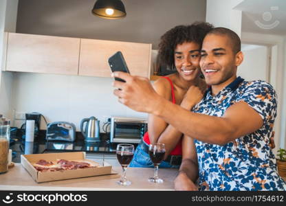 Portrait of happy young latin couple taking a selfie with mobile phone while having dinner at new home. Lifestyle and relationship concept.