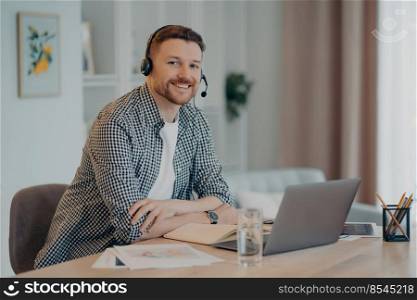 Portrait of happy young male freelancer sitting at his modern cozy workplace and working on laptop, cheerful guy smiling at camera and enjoying remote job. Freelance concept. Smiling man enjoying remote work at home, looking at camera
