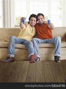 Portrait of happy young male friends holding tin cans while sitting comfortably on sofa at home