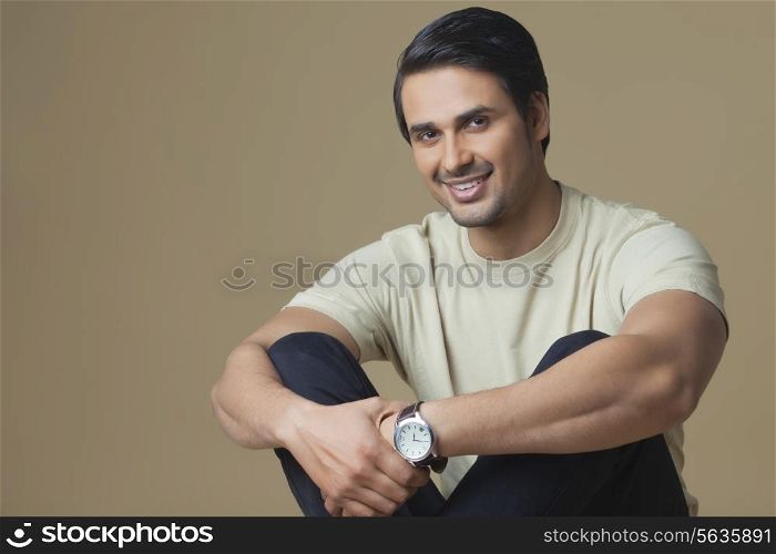 Portrait of happy young man hugging knees over colored background