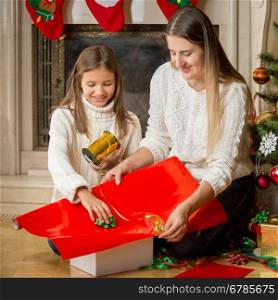 Portrait of happy young mother and cute daughter packing Christmas gifts in red paper and tying with golden ribbon