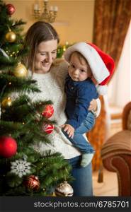 Portrait of happy young mother cuddling her baby boy in Santa cap at Christmas tree in living room