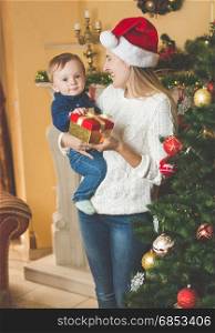 Portrait of happy young mother cuddling her baby boy in Santa cap at Christmas tree