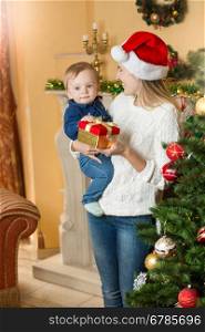 Portrait of happy young mother giving present to her baby son on Christmas at living room next to Christmas tree