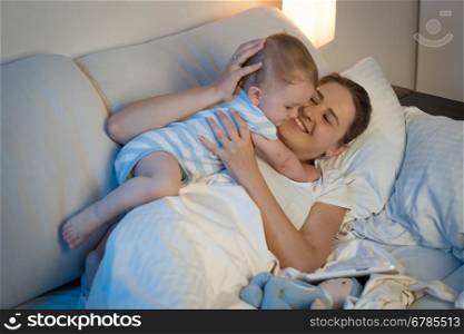 Portrait of happy young mother playing and embracing her baby boy in bed at late night