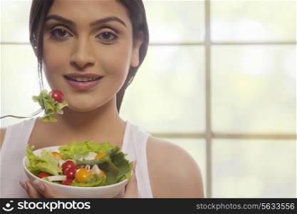 Portrait of happy young woman eating salad of lettuce, cherry tomatoes and mushrooms against glass window