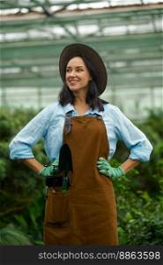 Portrait of happy young woman gardener entrepreneur wearing overalls proud of beauty of her own greenhouse. Young woman gardener proud of beauty of her own greenhouse