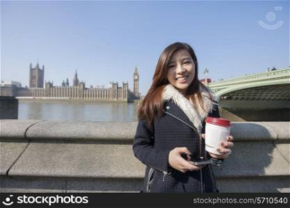 Portrait of happy young woman holding cell phone and disposable cup against Big Ben at London; England; UK
