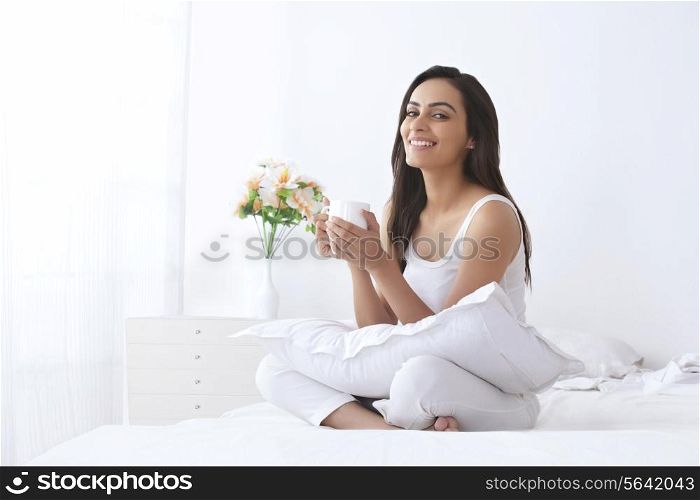 Portrait of happy young woman holding coffee cup on bed