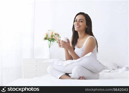 Portrait of happy young woman holding coffee cup on bed