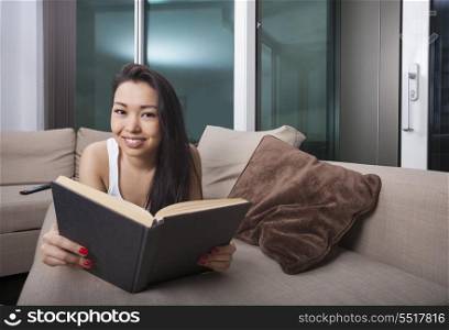 Portrait of happy young woman reading book while lying on sofa