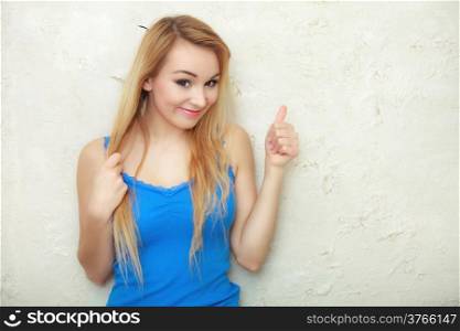 Portrait of happy young woman with long blond hair. Teenage girl showing thumb up success sign hand gesture. Blank copy space on white wall. Indoor.