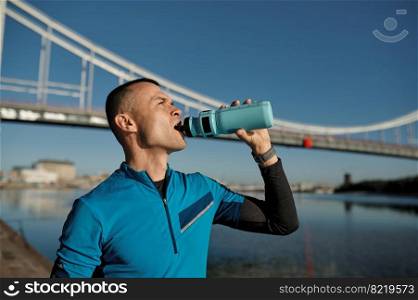 Portrait of healthy athletic middle aged man with fit body drinking water, resting after workout or running. Male runner drink after outdoor training against bridge. Portrait of athletic middle aged man holding water bottle for refreshing