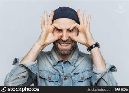Portrait of hppy bearded male makes eyewear with fingers, has stubble, wears jeans cloting, stands against white background. Funny middle aged man foolishes indoor, pretends wearing glasses.