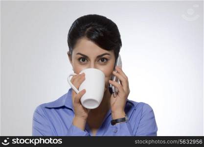 Portrait of Indian businesswoman drinking coffee while answering phone against gray background