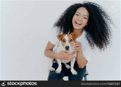 Portrait of joyful curly girl petting her dog, rejoicing buying jack russell terrier, smiles broadly, plays with animal, wears casual clothing, isolated over white background, enjoys good day