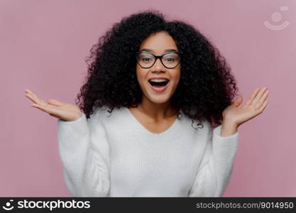 Portrait of joyful curly lady raises arms and spreads palms, wears optical glasses and white sweater, gets pleasant unexpected surprise from close person, isolated on violet studio wall. Happiness