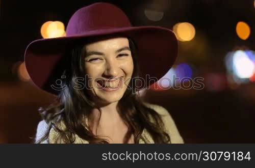 Portrait of joyful gorgeous female with long brown hair wearing stylish hat laughing as she stands on night city street. Positive emotional young woman looking at camera and smiling on summer night with coloful streetlights bokeh on background.