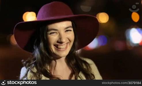 Portrait of joyful gorgeous female with long brown hair wearing stylish hat laughing as she stands on night city street. Positive emotional young woman looking at camera and smiling on summer night with coloful streetlights bokeh on background.