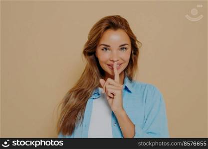 Portrait of joyful smiling female with long light brown hair holding index finger at her lips, saying &rsquo;shh&rsquo;, asking for silent or to keep her secret. Human face expressions, emotions and feelings. Portrait of joyful smiling female with long light brown hair