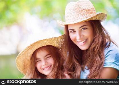 Portrait of joyful young mother with cute cheerful daughter wearing same straw hats and playing outdoors, laughing and looking on each other, portrait of a happy family enjoying life