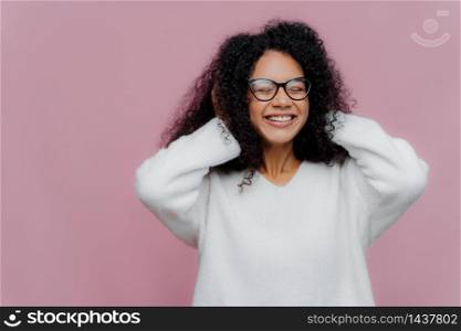 Portrait of joyous pleasant looking African American woman covers both ears with hands, hears loud music, smiles broadly, wears spectacles and white warm sweater, poses indoor, being very emotional