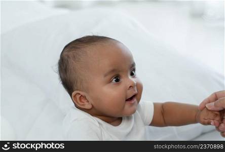 Portrait of little adorable African newborn baby sitting on bed and looking with bright eyes and smiling and holding mother hands at home. Motherhood, child care concept