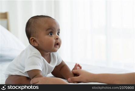 Portrait of little adorable African newborn baby sitting on bed and looking with bright eyes and smiling and holding mother hands at home. Motherhood, child care concept