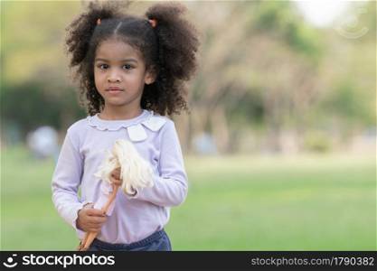 Portrait of little African kid girl with twin tails hair smiling and holding a doll in her hands at green park