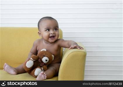 Portrait of Little African newborn baby girl wear diaper smiling and holding brown bear doll while sitting on yellow sofa at home. Innocent infant expression. White background