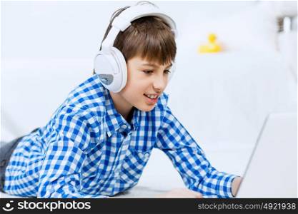 Portrait of little boy listening music from laptop through headphones while lying on hardwood floor. Boy of school age listening music from laptop through headphones while lying on floor