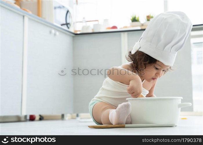 Portrait of little Caucasian adorable kid girl wear chef hat holding wooden spoon or whisk to stir food in pot. Cute child cooking and sitting on floor at kitchen at home