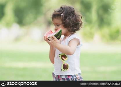 Portrait of little Caucasian cute girl holding sliced watermelon and enjoy eating or biting fresh fruit while picnic at park. Happy healthy child standing on green grass in summer