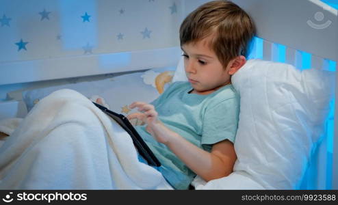 Portrait of little concentrated boy lying under blanket in bed and using tablet computer. Children with smartphones and gadgets at night.. Portrait of little concentrated boy lying under blanket in bed and using tablet computer. Children with smartphones and gadgets at night