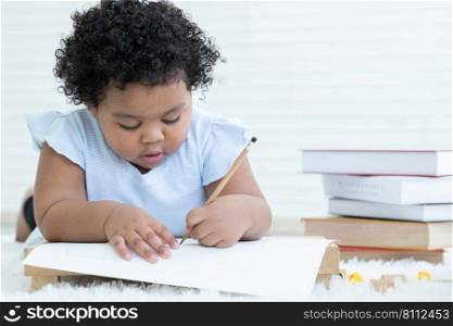 Portrait of Little cute African chubby kid girl use left hand writing on paper at table with stack of textbooks and toys lying on floor at home. Child education concept. White background. Copy space