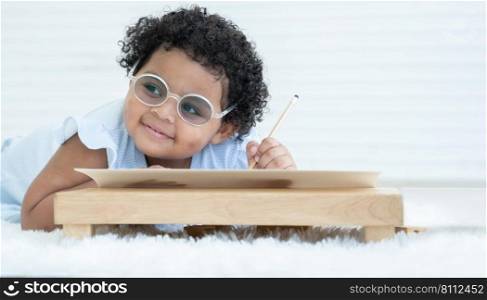Portrait of Little cute African chubby kid girl wear glasses smiling and use left hand writing on paper at table lying on floor at home. Child education concept. White background. Copy space
