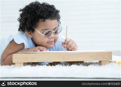 Portrait of Little cute African chubby kid girl wear glasses use left hand writing on paper at table lying on floor at home. Child education concept. White background. Copy space