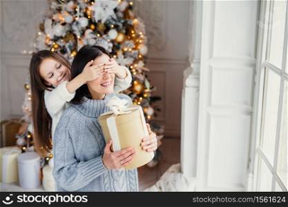 Portrait of little girl closes mother`s eyes, congratulates her with New Year or Christmas, stand near window in living room, have real miracle and feeling of holiday. Winter, celebration, season