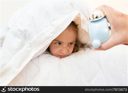 Portrait of little girl covering head with pillow and looking at alarm clock