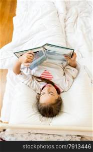 Portrait of little girl lying at bed in morning and reading big book