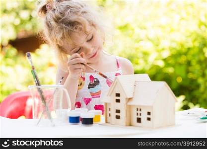 Portrait of little girl paints wooden model of house, summer outdoor, new home concept