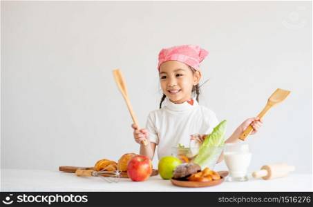 Portrait of little girl smile and holds wood ladle and fork in front of vegetables and fruits with concept of little chef on white background.