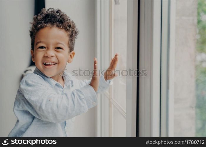 Portrait of little joyful kid with adorable smile and curly hair staying on windowsill next to closed large window, touching glass with small hands and smiling to camera in modern apartment interior. Portrait of little joyful black kid with adorable smile having fun at home