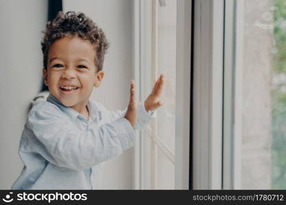 Portrait of little joyful kid with adorable smile and curly hair staying on windowsill next to closed large window, touching glass with small hands and smiling to camera in modern apartment interior. Portrait of little joyful black kid with adorable smile having fun at home