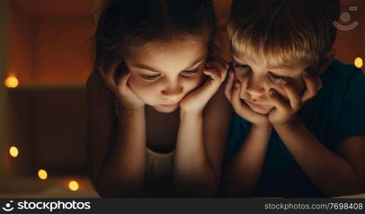 Portrait of little kids at night watching cartoons on the tablet, sad brother and sister bored at home