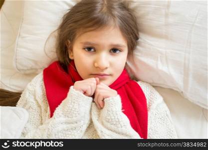 Portrait of little sad girl in white sweater lying under blanket at bed