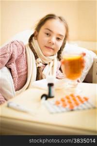 Portrait of little sick girl lying in bed taking cup of tea with lemon