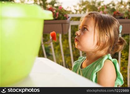 Portrait of little small girl blonde hair eating fruits watermelon in summer day by the table on her balcony