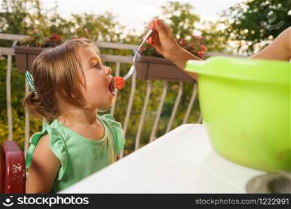 Portrait of little small girl blonde hair eating fruits watermelon in summer day by the table on her balcony fed by her mother